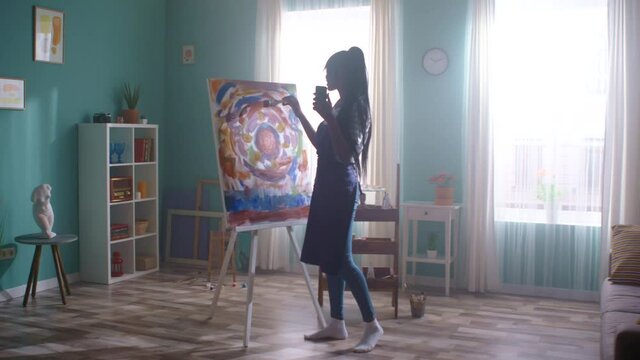 Beautiful African woman, blogger, is filming video on her gadget, spending weekend at creative studio, taking pleasure of hobby, painting on canvas with bright paints, Slow motion.