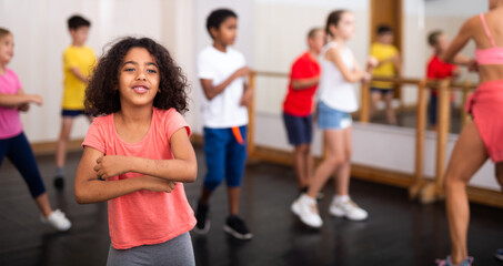 Portrait of cheerful little African American girl training movements during group class in dance school