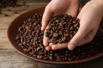 Woman taking pile of roasted coffee beans from bowl at wooden table, closeup