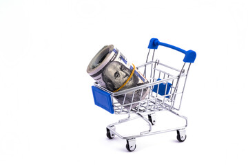 Rolled up 100 american dollar bills lie in a miniature supermarket shopping trolley on a white background