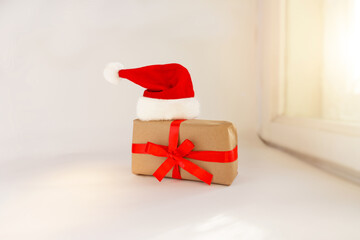 a gift box tied with a red ribbon with a Christmas hat lies on the window. The gift is wrapped in craft paper and red ribbon. Santa hat. Surprises and gifts for New Year and Christmas.
