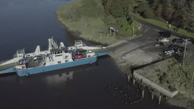 Compact blue river ferry approaches concrete riverside ramp, vehicles