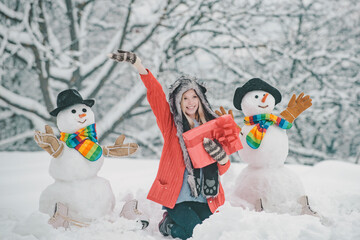 Girl playing with snowman in winter park. Snowman and funny young woman in winter hat and scarf.