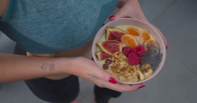 Millennial caucasian girl with tattoos in yoga outfit holding healthy fruit bowl. Diet breakfast. High angle, no face.