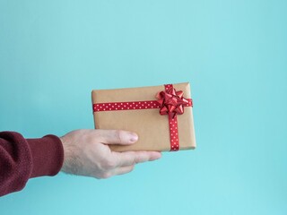 A man's hand holds a box with a gift with a red bow and satin ribbon on a blue background.