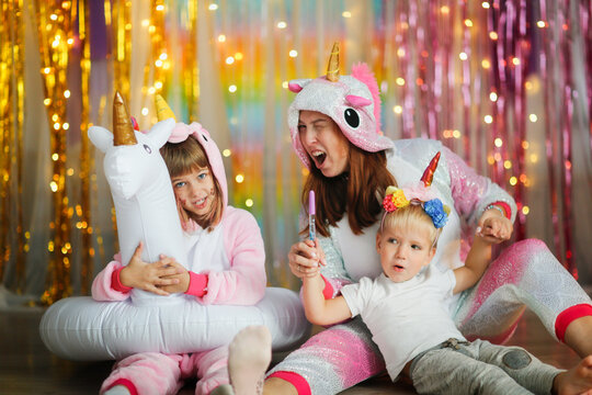 Funny Crazy Mom In A Unicorn Costume With Boy Toddler And Older Sister Together On Multicolored Rainbow Background With Lights With Unicorn, Children On Holiday In Style Of Unicorns, Photo Zone