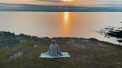 Aerial of young hipster woman sitting on blanket in front of Atlantic Ocean in France, Brittany taking photo during sunset golden hour. Incredible view after reaching top of Mountain. Wanderer.