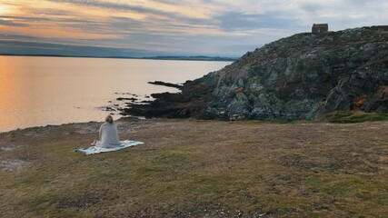 Beautiful young woman sitting and meditating on blanket on the top of mountain cliff in front of the Atlantic ocean at sunset. Female enjoying time at dreamlike Scenery during golden hour.