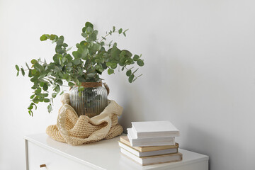 Vase with green eucalyptus branches and books on shelf near light wall