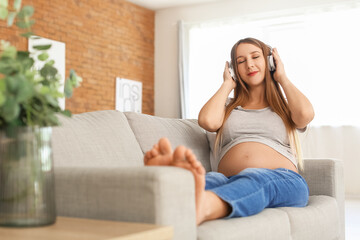 Young pregnant woman in headphones listening to music on sofa at home