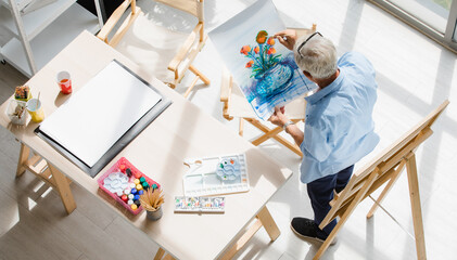Top view shot of Caucasian old senior elderly retired white bearded male painter wearing glasses sitting on wooden chair drawing painting picture with colorful watercolor with paintbrush on paper