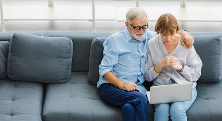 Caucasian old senior elderly lovely lover grandparents romantic couple gray hair and bearded husband wears glasses hugging wife shoulder sitting watching online show together on couch in living room