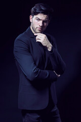 Portrait of Handsome Caucasian Brunet Businessman Wearing Black Suit Posing With Folded Hands And Touching Chin Against Black Studio Background.