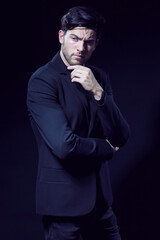 Fototapeta premium Portrait of Handsome Caucasian Brunet Businessman Wearing Black Suit Posing With Folded Hands And Touching Chin Against Black Studio Background.