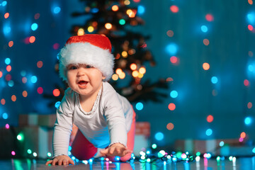 Happy little baby in a Christmas hat crawls near festive lights and xmas gifts. Indoor. Copy space. Winter seasonal holidays concept