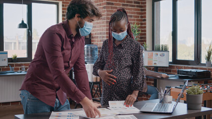 Pregnant employee working with man on business charts, doing teamwork to plan project strategy, wearing face masks. Woman expecting child and doing assignment at office during pandemic