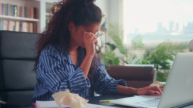 Overworked tired female student experiencing eye fatigue after working on laptop. Indian young woman with curly hair rubs her eyes and nose suffering from dry irritated eyes and bad blurry vision.