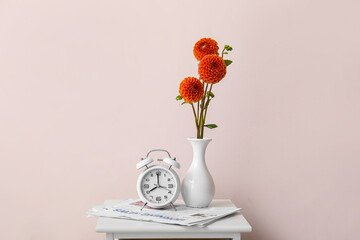 Vase with beautiful dahlias, alarm clock and newspaper on table near color wall