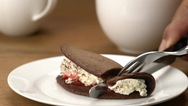 Using knife and fork cut dessert mini black forest pancake with fresh cream in white ceramic plate and take a bite to eat.