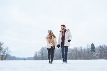 Young happy couple smile to each other and walk in winter park holding hands. Man and woman have fun together during winter holidays.