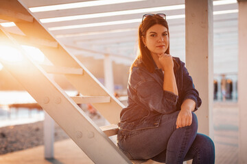 A young beautiful confident woman poses sitting on a white wooden staircase. Sunset in the background. The concept of confidence