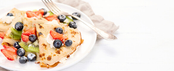 Sweet crepes with berries and cream on white wooden background.