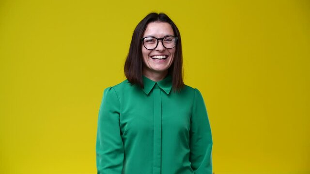 the girl looks at the camera and shows that you would call her, she is wearing glasses, dressed in a green shirt, short hair, brunette, the background is yellow
