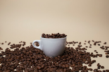 White Cup  full of coffee beans. With a lots of beans around. Light brown background.