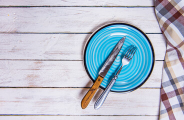 Top view background with empty blue ceramic plate, antique silver cutlery on vintage weathered white wooden boards. Copyspace. Brown checkered kitchen towel right corner.
