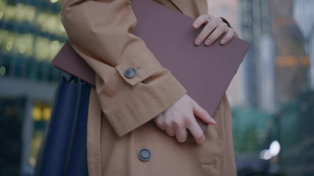 Office worker young woman wearing fancy coat goes from work holding laptop under arm against modern financial center and walking people in evening closeup