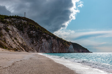 Epic stormy clouds on warm summer Mylos pebble beach. Idyllic coast with high cliffs of Lefkada island in Greece. Summer travel to turquoise Ionian Sea