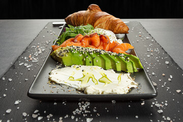 Italian panini sandwich, french croissant with salmon, avocado, rukkola and cream cheese on a rectangle plate. Luncheonette, snack plate Close up, top view