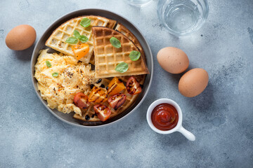 Plate of waffles served with scrambled eggs and tomatoes, flatlay on a light-blue stone background, horizontal shot
