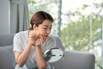 Upset stressed sad acne woman with problem skin squeezes pimple at home using a small round mirror