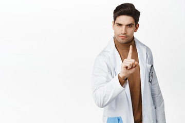 Image of doctor saying no, scolding, shaking finger in disapproval and looking skeptical at camera, standing over white background
