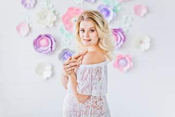 Smiling pregnant woman posing near white wall with decorative flowers. Portrait of young happy pregnant blonde in white interior. Happy motherhood.