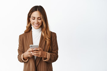 Smiling corporate woman order taxi in app, chatting in messanger app on smartphone, looking at her phone screen with pleased face, white background