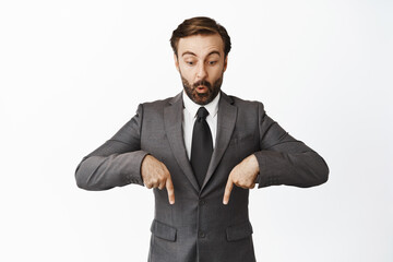 Surprised male entrepreneur pointing fingers down and looking below at advertisement, interesting promo offer, sale downwards, standing over white background