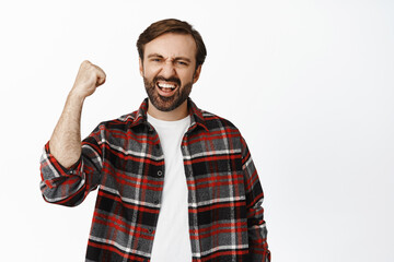 Portrait of enthusiastic bearded man raising clench fist and rooting for team, enjoying watching game, standing over white background