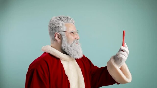 Modern Santa Claus. Cheerful Santa Claus is talking on the phone, by video call, at a distance, he says hello, wishes you merry christmas. blowing snow into telephone. blue background. slow motion
