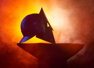 Iron helmet with face protection lying on anvil at forge. Smoke and fire on background. Traditional...