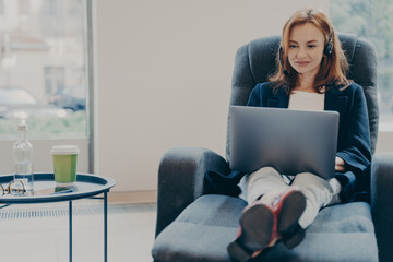 Fototapeta na wymiar Smiling red-haired woman working in headphones on laptop computer in modern open plan office