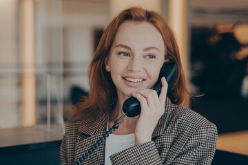 Portrait of satisfied female office worker using black landline phone, calling business partner - obrazy, fototapety, plakaty
