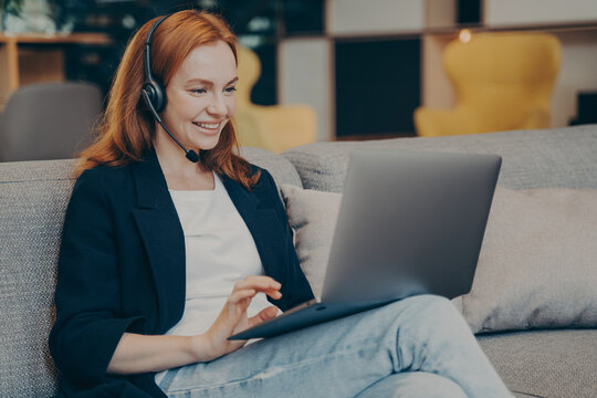 Young positive gingerhead female student having online webinar via laptop and headset in coffee shop
