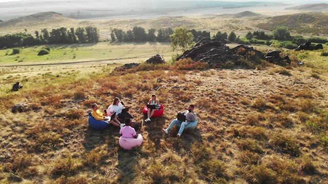 Five interracial freelancers sitting in soft chairs nature with laptops and coffee are having conversation, aerial view. Few friends sitting in chairs bags and talking. Concept of collaboration