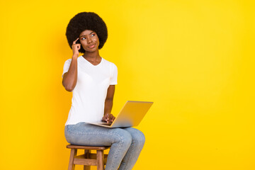 Photo of charming thoughtful dark skin lady wear t-shirt sitting chair typing device looking empty space isolated yellow color background