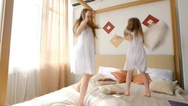Two sisters girls in white dresses jumping in the bed at home with daylight. Cute female siblings kids playing in the bedroom