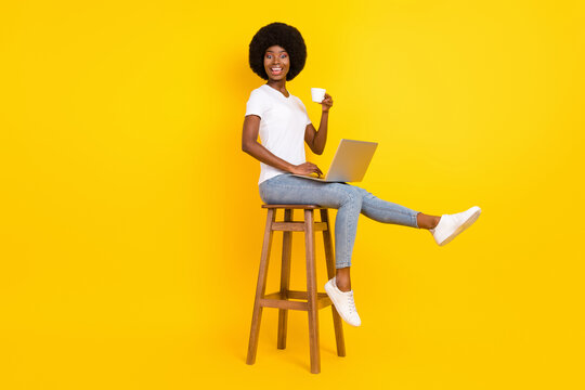 Photo of sweet charming dark skin lady wear t-shirt sitting bar chair typing modern device drinking tea isolated yellow color background