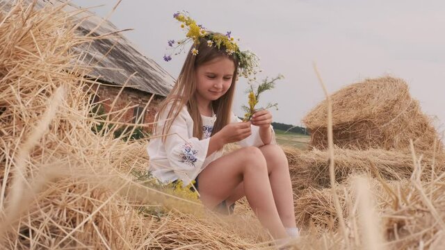 Ukrainian preteen girl wearing national dress and flower wreath sitting on hay stack. Pretty child in countryside in summertime. Female teen on farm outside with old house on background