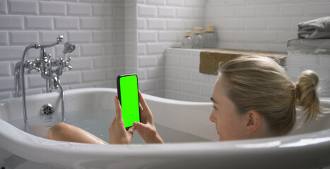 Beautiful Blonde Girl Takes a Bath Uses Smartphone with Green Mock-up Screen while Relaxing....